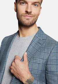 Kraciasty blezer w niebieskim, zielonym i białym kolorze, z klapami w kształcie litery V i dwoma guzikami. Noszony na szarym T-shircie, prezentującym nowoczesny krój.