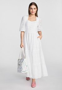 Biała bawełniana sukienka maxi z bufiastymi rękawami, ażurowymi detalami, kwadratowym dekoltem i zapięciem na guziki. Modelka trzyma torebkę w kwiatowy wzór.