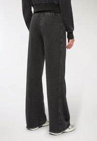 Osoba ubrana w luźne czarne spodnie i białe trampki stoi plecami do aparatu na białym tle.