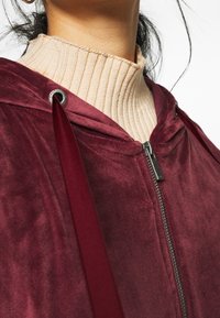 Sudadera de terciopelo burdeos con cremallera, detalles de cordón y un collar beige acanalado, con una textura suave y herrajes brillantes.
