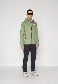 Light green waterproof jacket with a hood, zip front, and side pockets, paired with black pants and light-colored shoes.