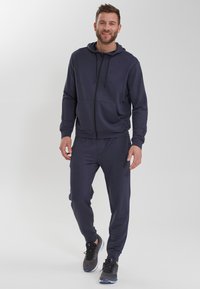 Mörkblå hoodie med dragsnören, dragkedja och framfickor, ihopparad med matchande sweatpants och svarta träningsskor. Slät tygtextur.