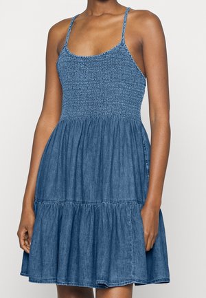 Kobieta w niebieskiej denimowej sukience bez rękawów, z cienkimi ramiączkami, marszczonym stanikiem i warstwową spódnicą, stoi na jednolitym tle.