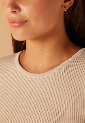 Geribbelde beige trui met een ronde halslijn, met een soepele textuur. Gouden oorbellen met kleine hangende details zichtbaar nabij het oor van het model.