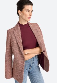 Blazer à carreaux marron et rouge avec revers en pointe, fermeture à un bouton, et poches, sur un top côtelé bordeaux, associé à un jean bleu clair.