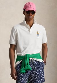 Mann iført hvit polo med teddybjørnlogo, marineblå shorts med palmetrær, grønn genser knyttet rundt livet, rosa caps og solbriller.