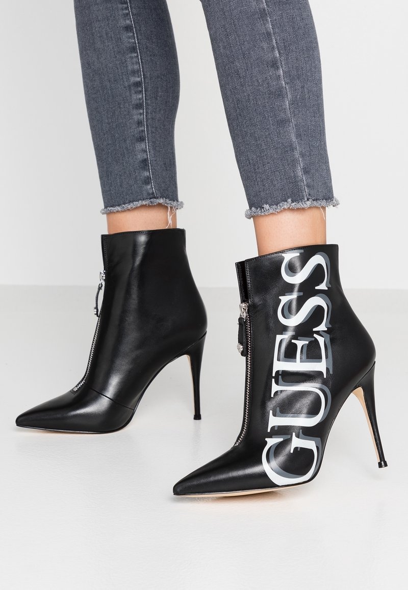 Bottes noires à talons aiguilles et bouts pointus avec fermetures éclair à l'avant et grandes lettres blanches "GUESS" sur les côtés extérieurs, portées avec un jean gris effiloché.