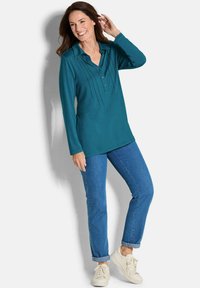 Teal-langärmeliges Polo-Shirt mit Knopfleiste und vertikalen Fältchen, kombiniert mit blauen Jeans mit umgeschlagenen Ärmeln und weißen Sneakern.