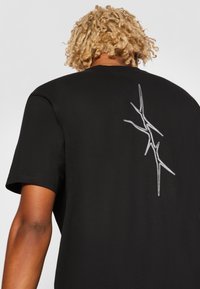 Black cotton t-shirt featuring a silver abstract design on the back. Short sleeves and crew neck style. Soft texture with a relaxed fit.