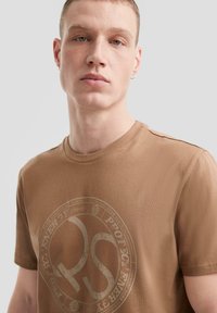 Camiseta de algodón marrón con cuello redondo y un gran estampado gráfico circular en el frente, que representa "RS" y elementos de texto.