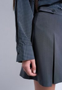 Femme portant une chemise à manches longues à carreaux foncés et une jupe gris foncé avec un pli à l'avant et une ceinture boutonnée.