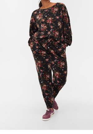 Kvinde iført en sort blomsterprintet langærmet top og matchende bukser med lommer, kombineret med lilla ruskindssneakers, stående med det ene ben krydset.