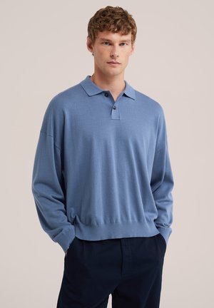 Giovane uomo con capelli ricci che indossa una polo blu a maniche lunghe larga e pantaloni scuri con le mani in tasca davanti a uno sfondo semplice.