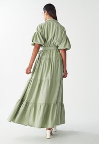 Svetlozelené maxi šaty s balónovými rukávmi, s tierovými volánmi a viazaním v páse. Hladká textúra a voľný dizajn, doplnené bielymi lodičkami.