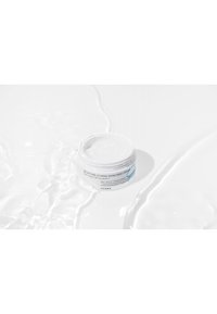 Crème enrichie en pouvoir hydratant dans un conteneur rond blanc avec un couvercle lisse, posée sur une surface blanche réfléchissante. Présente un design minimaliste.