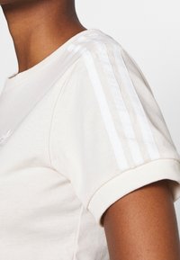 Světle béžové tričko s krátkým rukávem, žebrovanými manžetami a třemi bílými pruhy podél ramene. Měkká bavlněná látka s přiléhavým střihem.