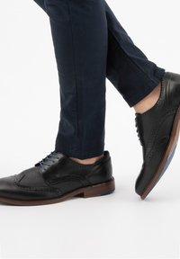 Scarpe eleganti in pelle nera con dettagli brogue, dotate di lacci blu e suola marrone. Indossate con pantaloni blu scuro, mostrando la vista laterale della scarpa.