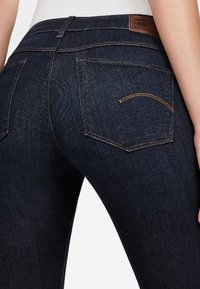 Jeans de mezclilla azul oscuro con una forma ajustada, con un parche de cuero marrón y dos bolsillos traseros con detalles de costura dorada.