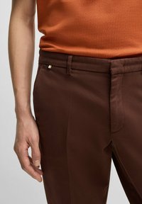 Mörkbruna byxor med en slät textur, med en bältesökning, platt front och en diskret metallisk detalj vid midjan.