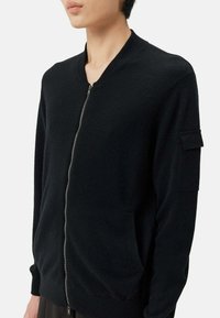 Svart zip-up jacka med ribbad krage, sidofickor och en ficka med lapp på ärmen, tillverkad av ett texturerat tyg.