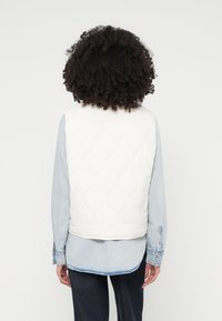 Bijela povučena prsluk s okruglim dijamantnim uzorkom, nošen preko svijetloplavog traper košulje. Stražnja strana prikazuje jednostavan, čist dizajn.