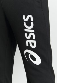 Černé tepláky s hladkým povrchem, na levé nohavici s velkým bílým logem "ASICS" tištěným vertikálně.