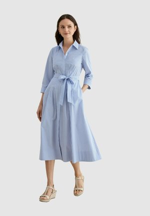 Mujer con cabello oscuro que lleva un vestido midi a rayas azules y blancas con mangas tres cuartos y cintura anudada, de pie con sandalias beige con cuña.