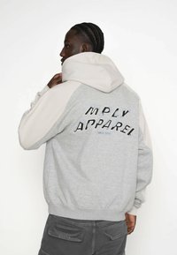 Šedá a béžová mikina s kapucí na šňůrku, text na zadní straně: "MPLY APPAREL SINCE 2015," a žebrované manžety a lem.