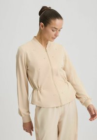 Veste bomber beige avec fermeture éclair à l'avant, taille froncée, manches longues et embellissements subtils. Tissu lisse avec poignets côtelés.