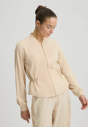 Giacca bomber beige con zip frontale, vita arricciata, maniche lunghe e sottili decorazioni. Tessuto liscio con polsini a costine.