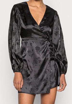 Schwarzes Wickelkleid aus Satin mit dezentem Blumenmuster, langen Puffärmeln, V-Ausschnitt und Taillenband an einer stehenden Person.