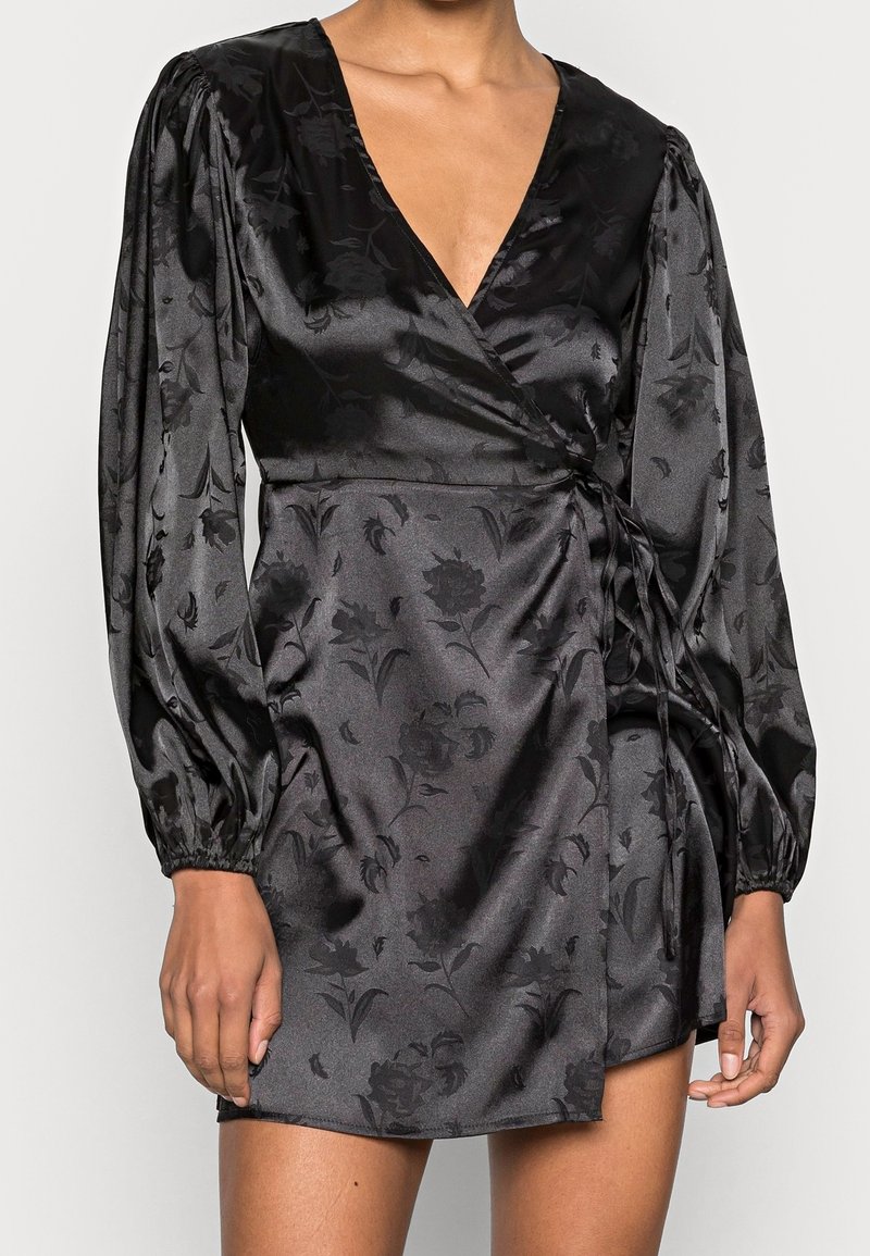 Robe portefeuille en satin noir avec un motif floral subtil, manches longues bouffantes, encolure en V et détail ceinture à la taille sur une personne debout.