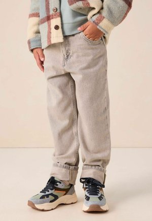 Child wearing light beige jeans rolled at the cuffs, colorful sneakers, and a multicolored plaid jacket with one hand in pocket.