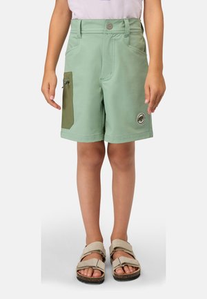 Bambino che indossa pantaloncini verde chiaro con una tasca laterale con zip e sandali beige, in piedi su uno sfondo uniforme.