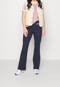 Person som bär en rosa kortärmad skjorta, beige vadderad väst, marinblå Adidas träningsbyxor och vita sneakers med blåa och röda accenter.