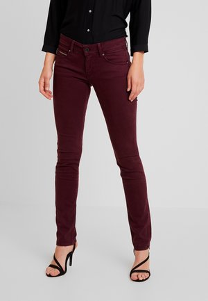Pantalones burdeos ajustados de algodón elástico con cinco bolsillos y cierre de cremallera. Combinados con tacones altos negros con tiras.