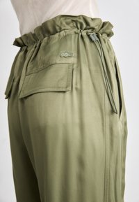 Light olive green trousers with an elastic waistband, side pockets, and a small embroidered logo. Smooth fabric with a relaxed fit.