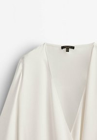 Blusa bianca a forma di involucro realizzata in tessuto morbido. Presenta una profonda scollatura a V e maniche lunghe con una texture liscia e fluida.