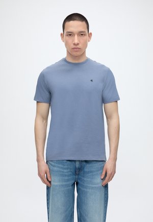 Mand iført en ensfarvet blå kortærmet T-shirt med et lille sort logo på brystet, kombineret med lyseblå jeans, står mod hvid baggrund.