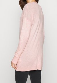 Světle růžový svetr s dlouhým rukávem, volným střihem, žebrovanými manžetami a mírně delším zadním lemem. Měkká textura, minimalistický design, bez viditelných vzorů.
