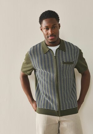 Mladý muž s krátkými kudrnatými vlasy, oblečený v olivově zelené krátkos rukávové pletené košili na zip s kostkovaným vzorem, stojící před jednobarevnou zdí.