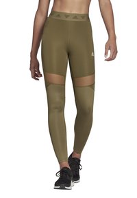 Olivengroene leggings met netuitsneden op de bovenbenen. Hoge elastische tailleband met een logo. Glad, rekbaar materiaal, gecombineerd met zwarte sportschoenen.