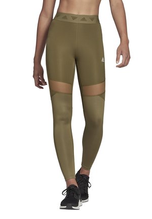 Olivengroene leggings met netuitsneden op de bovenbenen. Hoge elastische tailleband met een logo. Glad, rekbaar materiaal, gecombineerd met zwarte sportschoenen.