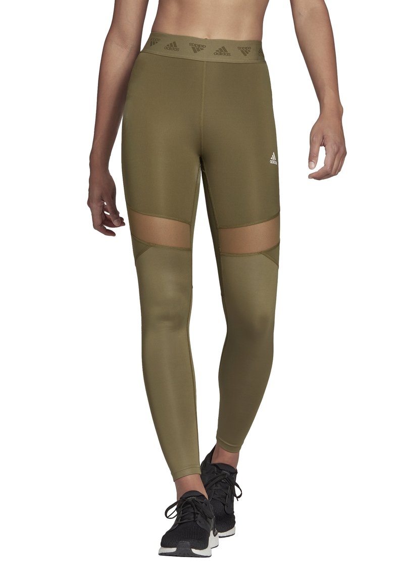 Olivengroene leggings met netuitsneden op de bovenbenen. Hoge elastische tailleband met een logo. Glad, rekbaar materiaal, gecombineerd met zwarte sportschoenen.