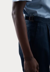 Granatowe spodnie w kant z teksturowanym wykończeniem, z metalowym zapięciem w talii i częściowo widoczną jasnoniebieską koszulą.