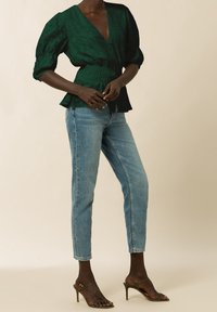 Blusa verde con texture, scollatura a V profonda, vita elastica e maniche a sbuffo abbinata a jeans attillati blu e tacchi a strappo.