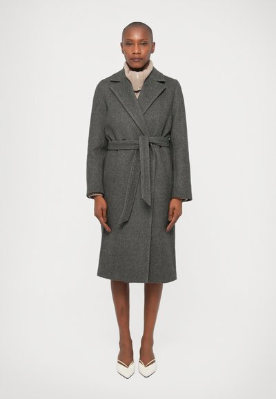 Cappotto in wool grigio con vita a cintura, collo a punte e maniche lunghe, abbinato a un dolcevita beige chiaro e scarpe con tacco alto bianche.