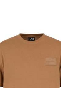 T-shirt in cotone marrone con colletto rotondo, maniche corte e una toppa testurizzata con dettagli del logo sul lato sinistro. Design semplice.