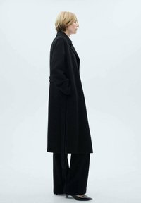 Palton lung negru cu un design structurat, realizat dintr-un material texturat. Dispune de un guler lat și buzunare laterale, asortat cu pantaloni evazați.