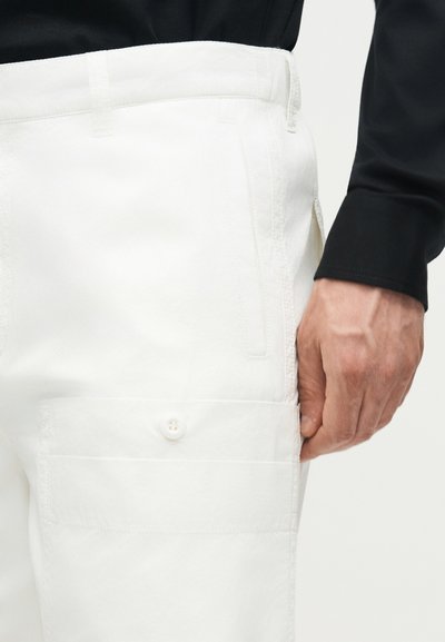 Ανδρικό χέρι που ακουμπά δίπλα στην τσέπη λευκού παντελονιού cargo με κλειστή με κουμπί πτέρυγα, φοράει μαύρο μακρυμάνικο πουκάμισο.
